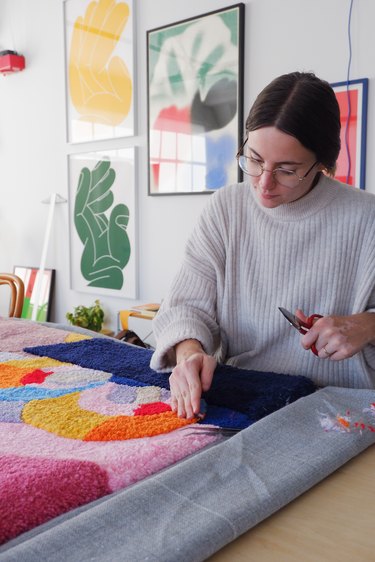 A dark-haired woman with her hair split down the middle and pulled back wears glasses and a white-ribbed turtleneck. She is holding scissors and working on a colorful patterned rug on the table.