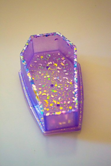 A translucent light purple coffin that doubles as a plant pot. It has multicolored star and moon glitter embedded in it.