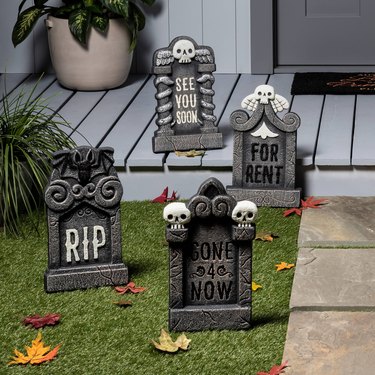 Four foam gray tombstones in a front yard and on a light blue-gray front porch. Three have skulls on them, while one has a bat with its wings open. Respectively, they say, "for rent," "see you soon," "RIP," and "gone 4 now."