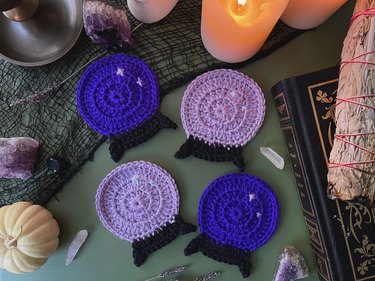 Four crocheted coasters in the shape of crystal balls. Two are light purple, while two are dark purple — all of them have two sparkles in the balls. They are on a dark sage green table with candles, a black and gold book, and amethyst crystals.