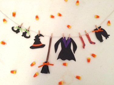 A yarn clothesline with cut-out witch clothes like pointy boots, a pointy hat, a long black and purple dress, a brown, striped orange and black socks, and black pantaloons with an orange bow. The item is surrounded by scattered candy corn.