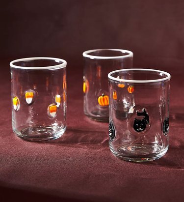 Three clear glasses, each with different little glass objects embedded in them. One has candy corn, one has plenty, and another has black cats. They are on a dark red backdrop.