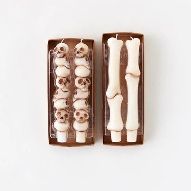 Two boxes. The one on the left contains two tapered candles shaped like stacked skulls. The one on the right how to candles shaped like bones sitting vertically on top of each other.