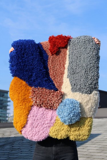 Multicolored patterned rug being held by two hands outside with blue sky  in the background