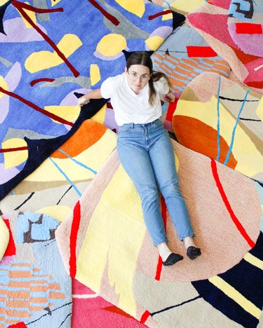 Woman with long dark hair and glasses wearing a white T-shirt, blue jeans, and black flats sits with legs outstretched on a layered and colorful patterned rugs.