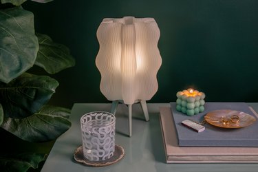 A white, wavy transparent, 3D-printed lamp on a shiny table next to a lush plant.