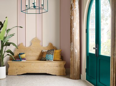 Wooden bench with multi-colored pillows in front of a baby pink wall next to a green-blue door