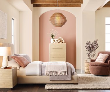 Pastel colored bed facing sideways in front of a beige and pink wall with a dresser, side table, and hanging light fixture. There is a rust-colored chair in the corner with a pink pillow next to a plant.