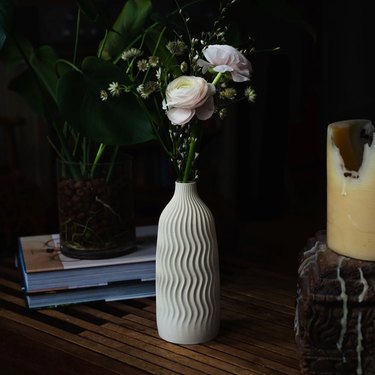 A wavy, 3D-printed white vase holding light pink flowers. The vase is on a medium-colored wood table with two stacked books and a melting candle.