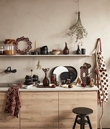 Kitchenware like plates, vases, and jars on a countertop with wooden shelf, a black stool, and an apron hanging on the beige wall.