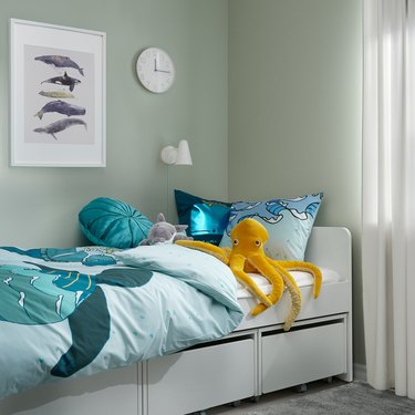 A children's bedroom with a white bed frame, a blue blanket with a turtle on it, blue pillows, and an orange octopus plush resting on the bed.