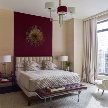 Cream bedroom with maroon accent wall, lamp, and bench.