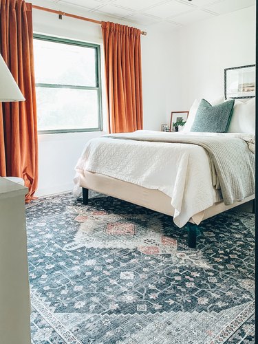 Bedroom with orange curtains, blue rug, and white bedding.