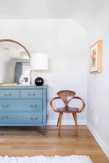 A white bedroom with a blue dresser with a large mirror, black lamp, and wooden chair next to it.