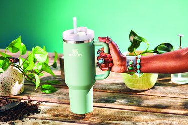 A Eucalyptus 40oz Stanley Quencher Tumbler is being held by a Black woman’s hand over a wooden surface filled with plants. The tumbler is light green with a white top, and a clear reusable straw, and the arm and hand is adorned with turquoise jewelry.