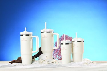 Four cream colored Stanley Quencher Tumblers rest on a small pile of sand amidst starfish, a sand dollar, a piece of driftwood and a piece of coral. The wall behind them are blue, and each size Tumbler is being shown, 30oz, 40oz, 14oz and 20oz.