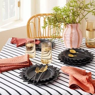 table with striped tablecloth, black dishes, gold silveware, striped vase with greenery, and orange-red napkins