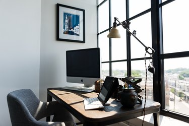 A wood desk with a computer and industrial lamp. A gray organic shaped chair. A large window with a black pane.