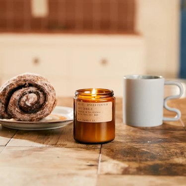 A spiced pumpkin candle in an amber jar on a wood table next to a white mug with a wavy handle and a dark brown cinnamon roll.