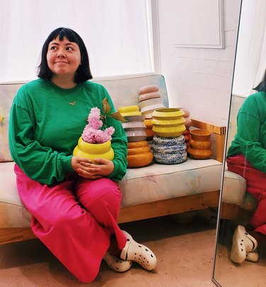 photo of a figure holding ceramics on couch next to pile of colorful ceramics and mirror nearby