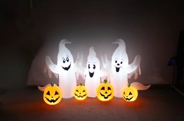 Three white ghosts with arms above their heads and four jack-o-lanterns as inflatable decor in a dark front yard.