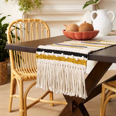table with fringed runner and orange bowl with fruit and white pitcher nearby
