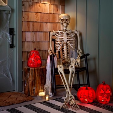 A fake skeleton sitting on a dark wood rocking chair on a front porch with light-up bright orange fake jack-'o-lanterns.