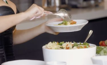 Kylie Jenner scooping pasta with vegetables from a large bowl and into a small white serving bowl