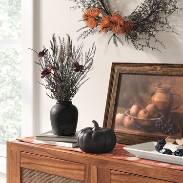 A black ceramic pumpkin on a wood storage table next to a black vase with black and purple dried florals in it.