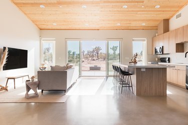 Kitchen and living area with island, bar stools, couch, tv, end table, bench, cabinets, windows, wood ceiling.