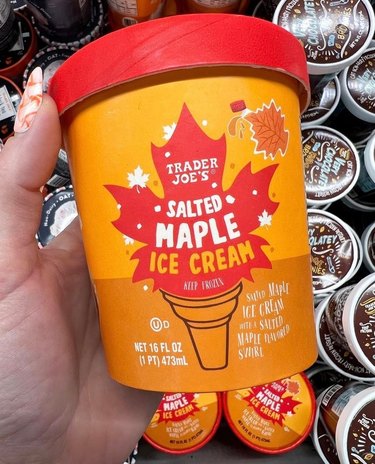 A pint of Trader Joe's Salted Maple Ice Cream is being held over the ice cream section of the freezer aisle in Trader Joe's. The carton is orange with a red top, and there's an image of a red maple leaf resting in an ice cream cone. There's a smaller orange maple leaf in the top right corner, and small white maple leaves throughout.