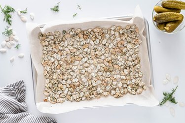 A parchment-lined silver baking sheet covered in the dill pumpkin seeds on a white counter. There is also a small bowl of dill pickles, a blue and white striped tea towel, and fresh dill and pumpkin seeds sprinkled on the white counter.