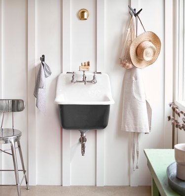 A black cast-iron wall-mounted sink on a white wall with paneling; a hat and an apron are hung near it