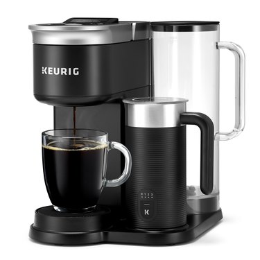 Keurig's newest brewer, the K-Cafe SMART is black with a silver top, and silver Keurig branding across the front of the brewer. The water reservoir is clear with a black top, and the milk frother is black with a silver top and a black handle. Coffee is being brewed into a clear coffee mug.