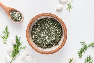 A small wood bowl filled with dried dill, sea salt, garlic powder, onion powder, and ground black pepper on a white counter.