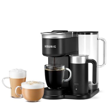 Keurig's newest brewer, the K-Cafe SMART, is a black and silver coffee brewer, with a black and silver milk brother attached. A clear coffee mug is catching brewed coffee, while two other clear mugs filled with lattes are to the side.