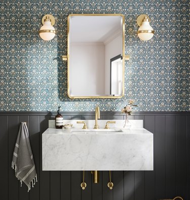 A marble wall-mounted sink with gold faucets and a gold trim mirror and sconces; a green patterned wallpaper and black wainscoting are on the wall