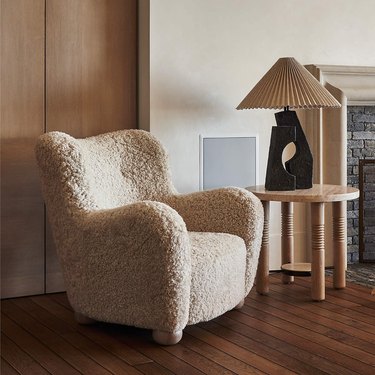 Fuzzy beige accent chair in a room on a wooden floor next to a wooden table with a beige lamp