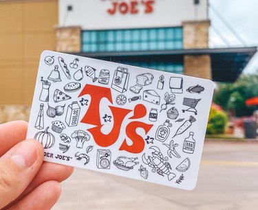 Hand holding up a Trader Joe's gift card in front of a store with the logo seen in the back.