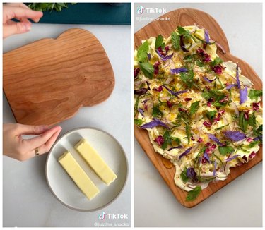 Hands with a wooden cutting board next to two sticks of butter on a white plate. On the right is a butter board garnished with herbs, onions, and edible flowers on a white countertop.