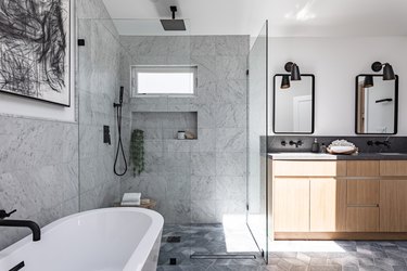 A gray bathroom with a glass shower door, a freestanding tub, and a light wood vanity with two mirrors