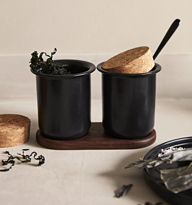 Two black cups on a wooden platform on a countertop with cork stoppers and silverware sticking out.