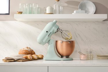 KitchenAid Blossom stand mixer in a soft green with a bronze bowl and gold mixer on a white countertop with white shelving with glasses and plates. There is also a load of bread on a wooden cutting board next to the mixer.