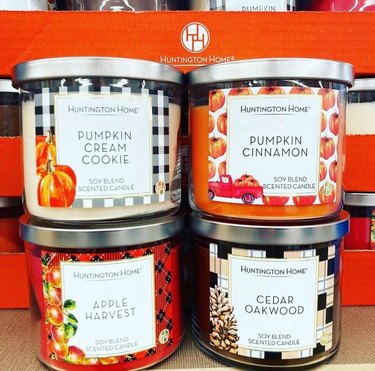 A close up of four seasonal Aldi candles. The Pumpkin Cream Cookie label has a black gingham background and pumpkins in the corner. The Pumpkin Cinnamon label has a pumpkin printed label with a small red truck in the corner. Apple Harvest has a red plaid label with apples on a branch on the left side, and Cedar Oakwood has a tan, white and black checkerboard label with a pinecone pictured in the bottom left corner.