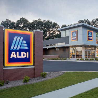 Exterior shot of an Aldi grocery store. An Aldi sign is prominent, with the Aldi store front behind it.