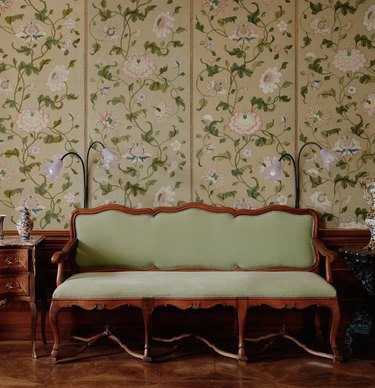 Two floral lamps with two stems and lavender heads sandwiching a wooden bench with green cushions in front of floral wallpaper