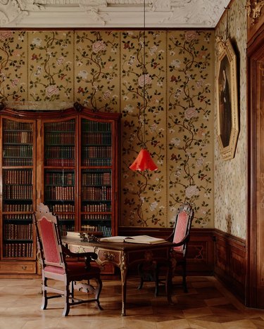 A red, floral-shaped hanging light over a wooden desk with two wooden chairs with red cushions. There is a bookcase in the background leaning against floral wallpaper.