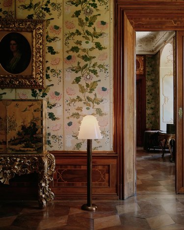A lamp with a curved head on a wooden floor in front of floral wallpaper, a wooden doorway, and antique picture frames on the wall