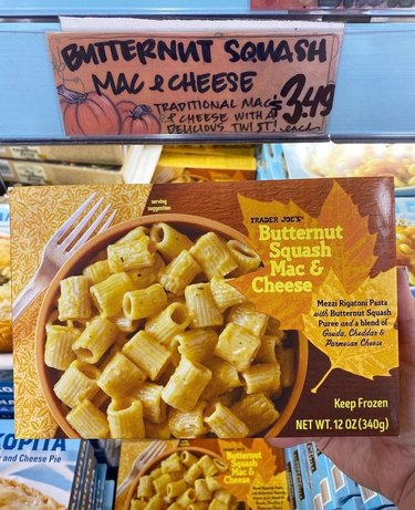 A box of Trader Joe's Butternut Squash Mac and Cheese is being held over the freezer section of a Trader Joe's aisle. The box has an orange bowl of rigatoni pasta, covered in squash puree and a three cheese blend with a fork resting next to it on the left. There is an image of a dark orange maple leaf with yellow text reading "Trader Joe's Butternut Squash Mac and Cheese" and a light orange leaf with text on it that reads "Mezzi rigatoni pasta with butternut squash puree and a blend of gouda, cheddar and parmesan cheese".