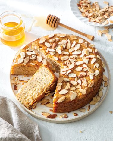 Honey-almond cake with a slice missing on a white plate sitting on a white table. There is a jar of honey and a plate of sliced almonds in the background.